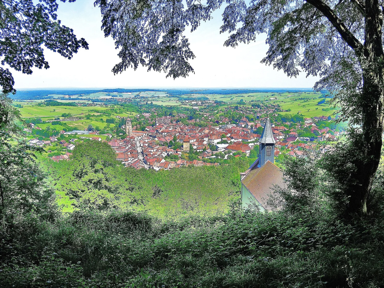 Vue plongeante sur les toits rouges d’Arbois et un clocher, encadrée de feuillages, avec la campagne verte qui s’étend jusqu’à l’horizon.