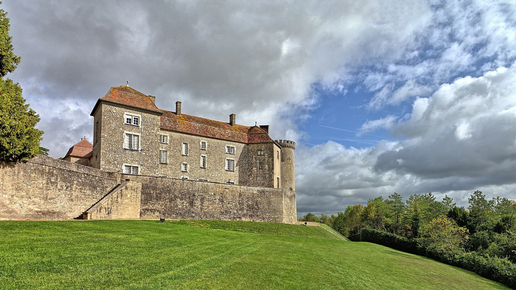 Château de pierre à toit de tuiles brunes flanqué d’une tour ronde, au sommet d’une pelouse en pente bordée d’un mur, sous un ciel nuageux.