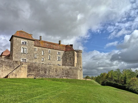 Château de pierre à toit de tuiles brunes flanqué d’une tour ronde, au sommet d’une pelouse en pente bordée d’un mur, sous un ciel nuageux.