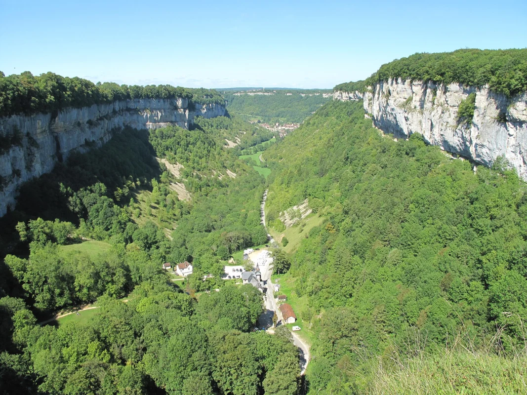 Vallée profonde et boisée encadrée de hautes falaises calcaires grises, avec quelques bâtiments et une route au fond, sous un ciel bleu.
