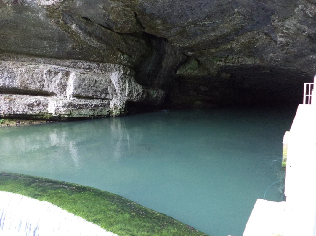 Vasque d’eau turquoise au pied d’une large grotte creusée dans une falaise calcaire, avec un muret et un garde-corps au premier plan.