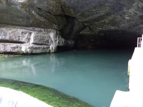 Vasque d’eau turquoise au pied d’une large grotte creusée dans une falaise calcaire, avec un muret et un garde-corps au premier plan.