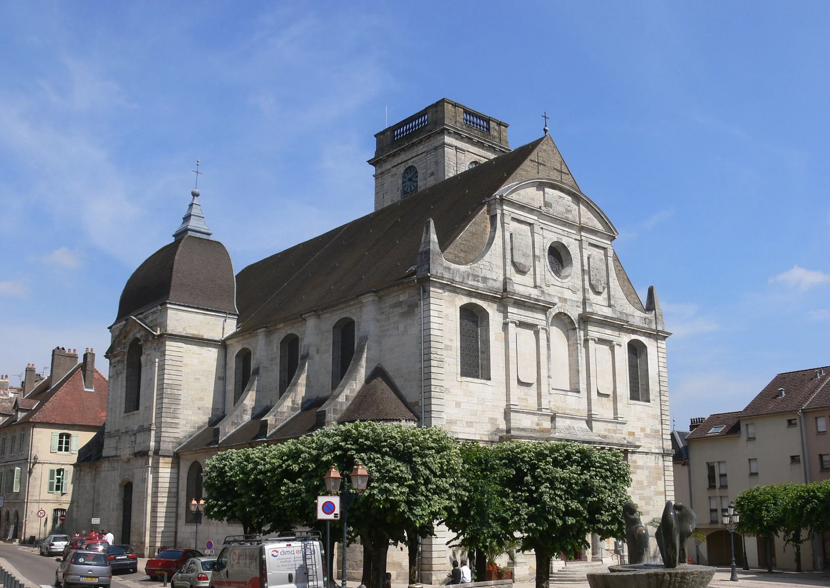 Église en pierre claire avec une tour carrée à toit plat et un dôme d’ardoise, devant une rue bordée d’arbres taillés sous un ciel bleu.