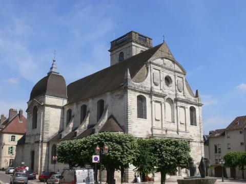 Église en pierre claire avec une tour carrée à toit plat et un dôme d’ardoise, devant une rue bordée d’arbres taillés sous un ciel bleu.
