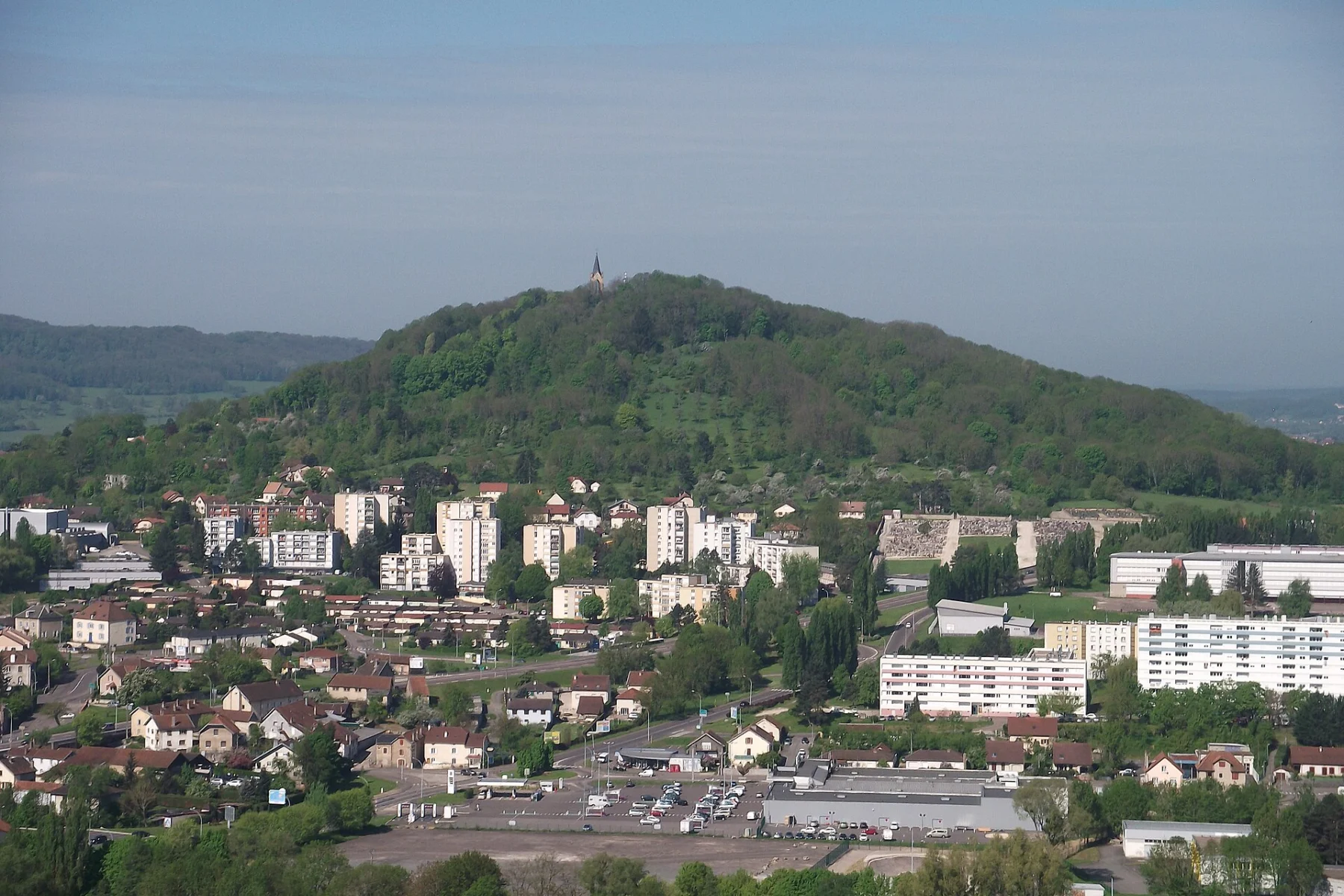 Colline boisée arrondie surmontée d’une petite chapelle claire, dominant un quartier d’immeubles et de maisons, avec des collines bleutées à l’arrière-plan.
