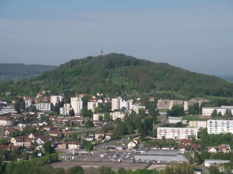 Colline boisée arrondie surmontée d’une petite chapelle claire, dominant un quartier d’immeubles et de maisons, avec des collines bleutées à l’arrière-plan.
