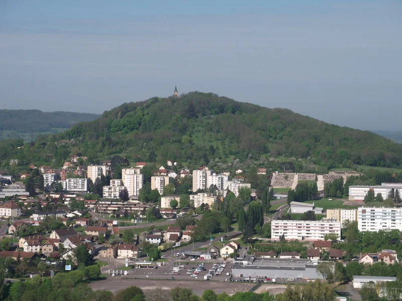 Colline boisée arrondie surmontée d’une petite chapelle claire, dominant un quartier d’immeubles et de maisons, avec des collines bleutées à l’arrière-plan.