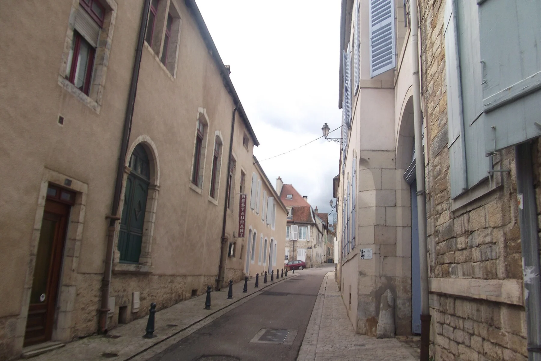 Rue étroite bordée de façades anciennes en pierre claire, aux fenêtres hautes et volets clairs, qui file entre deux rangées de maisons sous un ciel blanc.