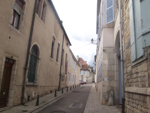 Rue étroite bordée de façades anciennes en pierre claire, aux fenêtres hautes et volets clairs, qui file entre deux rangées de maisons sous un ciel blanc.