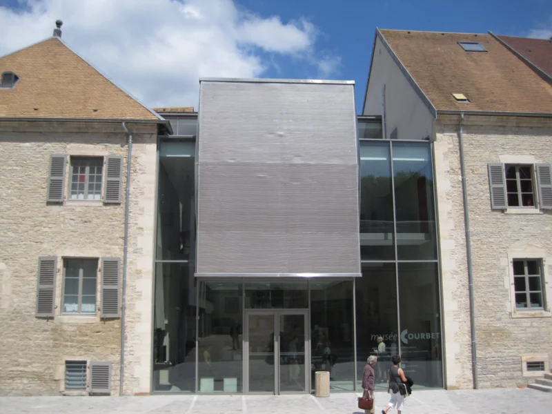 Façade contemporaine vitrée insérée entre deux maisons anciennes en pierre, entrée d'un musée.
