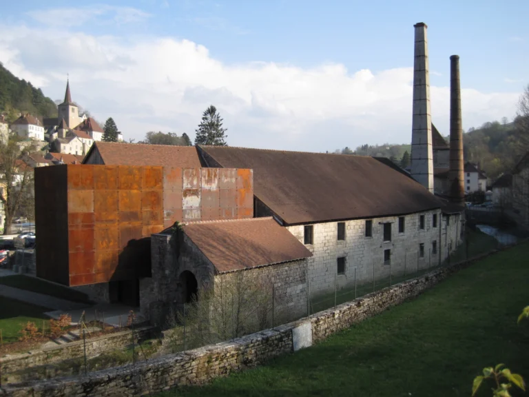 Bâtiments industriels anciens aux toits de tuiles, avec deux hautes cheminées en brique et un bâtiment moderne en acier rouillé.
